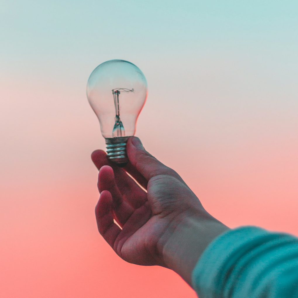 Hand holding a glowing lightbulb against a pink and teal sky